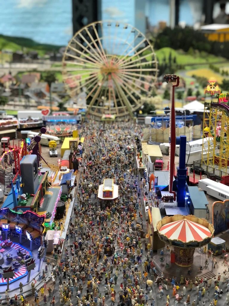 zentral ein Riesenrad und links und rechts Stände und Fahrgeschäfte eines Rummels, dazu viele Personen, alles als Modell